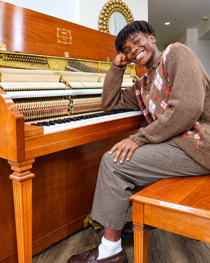 Justin Abraham Paul apoyado en su piano vertical, sonriendo